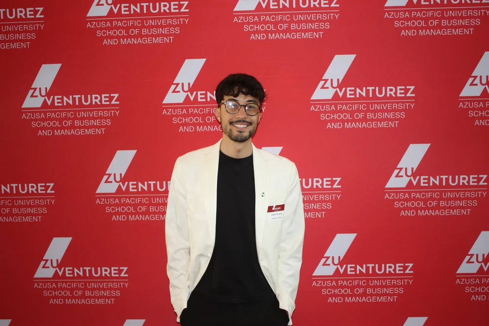 Julián Orozco con chaqueta blanca delante del photocall rojo de ZuVenturez, la competición de emprendimiento de la Azusa Pacific University.